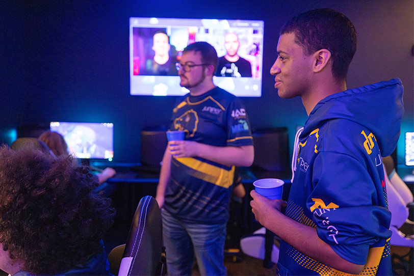Two people wearing esports jerseys, one in a blue and yellow shirt and the other in a blue and yellow hoodie, are holding cups and watching a screen displaying other people in a dimly lit gaming room. A third person with curly hair is visible in the foreground, seated and facing a computer monitor.
